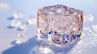 Melting ice cube with water drops on light blue surface, crystal clear frozen block catching sunlight with sparkling reflections and refractions.