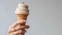 Creamy soft serve ice cream in waffle cone held by female hand with gold ring against neutral gray background. Perfect for food advertising.