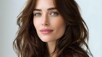 Young Caucasian woman with long dark brown hair, blue eyes and natural makeup looking at camera with gentle smile against light background. Professional beauty portrait.