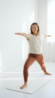 Young Caucasian woman practicing yoga warrior pose on mat in bright white studio room with natural light streaming through windows for wellness and fitness.