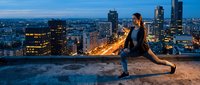 Athletic woman stretching on urban rooftop at dusk with illuminated city skyline and glowing street lights creating dramatic evening fitness scene.