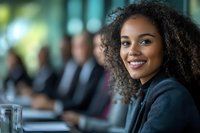 Confident young African American businesswoman with curly hair smiling during corporate meeting with colleagues in background, professional workplace setting.