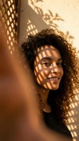 Young mixed-race woman with curly hair smiling in warm sunlight with geometric shadow patterns from window lattice creating artistic lighting effects on face.