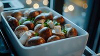 Fresh mushrooms with mozzarella cubes and herbs in white ceramic dish, moody lighting with bokeh background creates appetizing composition for food photography.