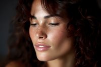 Close up portrait of young woman with closed eyes, natural makeup, and curly dark hair against dark background, showing peaceful serene expression.