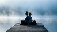 Two people practicing meditation yoga on wooden dock over misty lake at dawn creating peaceful zen atmosphere for wellness and mindfulness retreat.
