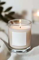 Lit aromatic candle in clear glass container with blank white label on soft fabric, creating cozy atmosphere with gentle flame and warm glow.