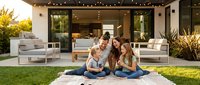 Young family with two children enjoying quality time on lawn in front of modern home with contemporary outdoor furniture and large glass doors, representing residential lifestyle and togetherness.