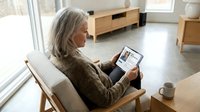 Senior woman with gray hair reading news on tablet computer while sitting in modern living room chair with coffee cup nearby.