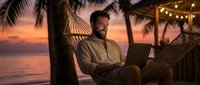 Happy bearded man working on laptop in hammock at tropical beach sunset with palm trees and string lights for remote work vacation concept.