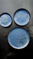 Artisanal ceramic plates in dusty blue glaze with speckled texture and rustic bronze rim on dark background, arranged in minimalist composition for tableware styling.