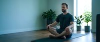 Relaxed middle-aged Caucasian man sitting cross-legged on yoga mat in modern living room with plants by window for meditation and mindfulness practice.