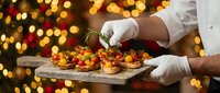 Chef hands in white gloves presenting gourmet appetizers on wooden board with festive bokeh lights background for holiday catering and fine dining events.