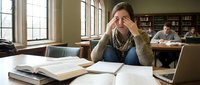 Stressed young Caucasian female student sitting in library with open books and laptop, holding head in hands, struggling with studies.