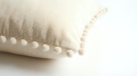 Beige linen throw pillow with decorative pom pom trim on white background, cozy boho home decor accent for living room or bedroom styling.