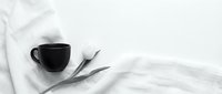 Black ceramic coffee cup and white tulip flower on soft linen fabric, monochrome flat lay with copy space for text.