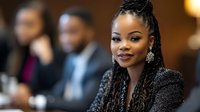 Professional African American businesswoman with braided hairstyle smiling confidently in corporate meeting environment with colleagues in background.
