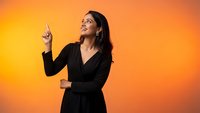 Young Hispanic businesswoman in black blazer pointing up with finger against orange gradient background for corporate presentations and marketing.