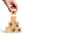 Hand placing wooden cube with lightbulb icon on pyramid of blocks featuring environmental symbols for sustainable business concepts.