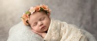 Peaceful newborn baby girl sleeping wrapped in cream lace blanket wearing delicate peach rose flower crown headband on soft textured backdrop.