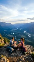 Young couple enjoying sunset picnic on mountain summit overlooking valley with winding river and dramatic alpine landscape during golden hour hiking adventure.