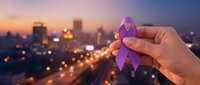 Purple ribbon held in hand against illuminated city skyline at dusk, symbolizing awareness and support for health causes and charitable initiatives.