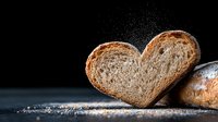 Heart-shaped slice of rustic bread with flour dust on dark background, dramatic lighting creates atmospheric mood for food photography and bakery marketing.