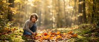 Young boy with light skin tone and brown hair, wearing casual clothing, crouching among fallen autumn leaves in a sunlit forest. Suitable for editorial, lifestyle, and seasonal content.