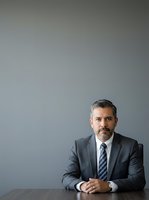 Middle-aged Caucasian male professional wearing dark gray business suit and striped tie, seated at desk with hands clasped, neutral gray background, suitable for corporate portraits, business websites, and professional headshots.