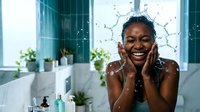 Young woman with dark skin tone splashing water on her face in a modern bathroom, expressing joy and freshness during skincare routine.