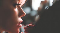 Close-up profile of young woman applying lipstick in soft lighting, creating intimate beauty moment with blurred background.