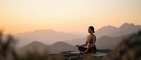 Young woman practicing meditation yoga pose on mountain cliff during golden sunset with layered peaks creating peaceful mindfulness wellness scene.