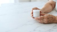 Senior woman with age spots and wrinkles holding white cosmetic jar on marble surface, showcasing skincare product for anti-aging beauty and wellness marketing.