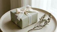 Elegant sage green gift box with cream ribbon bow on beige plate, dried branch beside it, soft natural light, minimalist style.