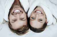 Young couple lying head to head in white bathrobes, smiling with bright teeth and relaxed expressions during spa treatment.