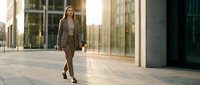 Young woman with blonde hair wearing brown business suit walking through modern urban corridor with glass and concrete architecture during daytime.