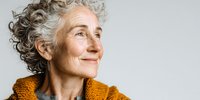 Senior woman with curly gray hair and warm smile wearing orange sweater against white background, portraying confidence and contentment in aging.