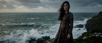 Young woman with long dark hair in floral dress standing on dramatic rocky coastline with crashing ocean waves and stormy sky for fashion portrait photography.