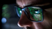 Close-up of programmer wearing glasses with binary code reflection, dark environment with green digital data stream showing cybersecurity concept.