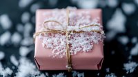 Pink wrapped present box with gold twine bow dusted in white snow crystals against dark background, creating magical winter holiday atmosphere.