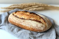 Freshly baked artisanal sourdough bread loaf with crispy crust and distinctive scoring pattern on gray linen cloth, decorated with wheat stalks in background.