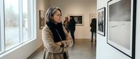 Mature woman with gray hair contemplating artwork in modern gallery space with natural lighting and framed photographs on walls.