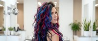 Woman with long wavy hair dyed in vibrant blue and red streaks, standing in modern salon interior with mirrors and plants, showcasing bold hair color transformation and styling.