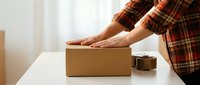 Person in plaid shirt packing cardboard box on white table with tape dispenser, preparing shipment or moving supplies for delivery and logistics.