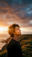 Young man with curly hair meditating peacefully at sunset with dramatic orange and blue sky clouds creating serene outdoor wellness moment.