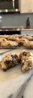Freshly baked chocolate chip cookie broken in half showing gooey melted chocolate chunks against marble counter, with cooling rack of cookies in background.