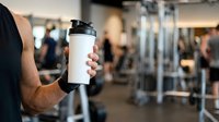 Athlete holding blank white water bottle in gym with blurred fitness equipment and people in background, suitable for beverage branding and sports marketing.