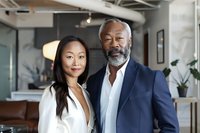 Asian woman in white blazer and African American senior businessman with gray beard smiling in modern office interior with industrial design elements.