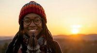 Young African American woman with dreadlocks wearing knit beanie and glasses smiling at golden hour sunset outdoors for lifestyle and wellness content.