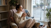 Young woman relaxing at home with hot beverage, sitting in cozy armchair by window. Comfortable indoor lifestyle moment with natural lighting.